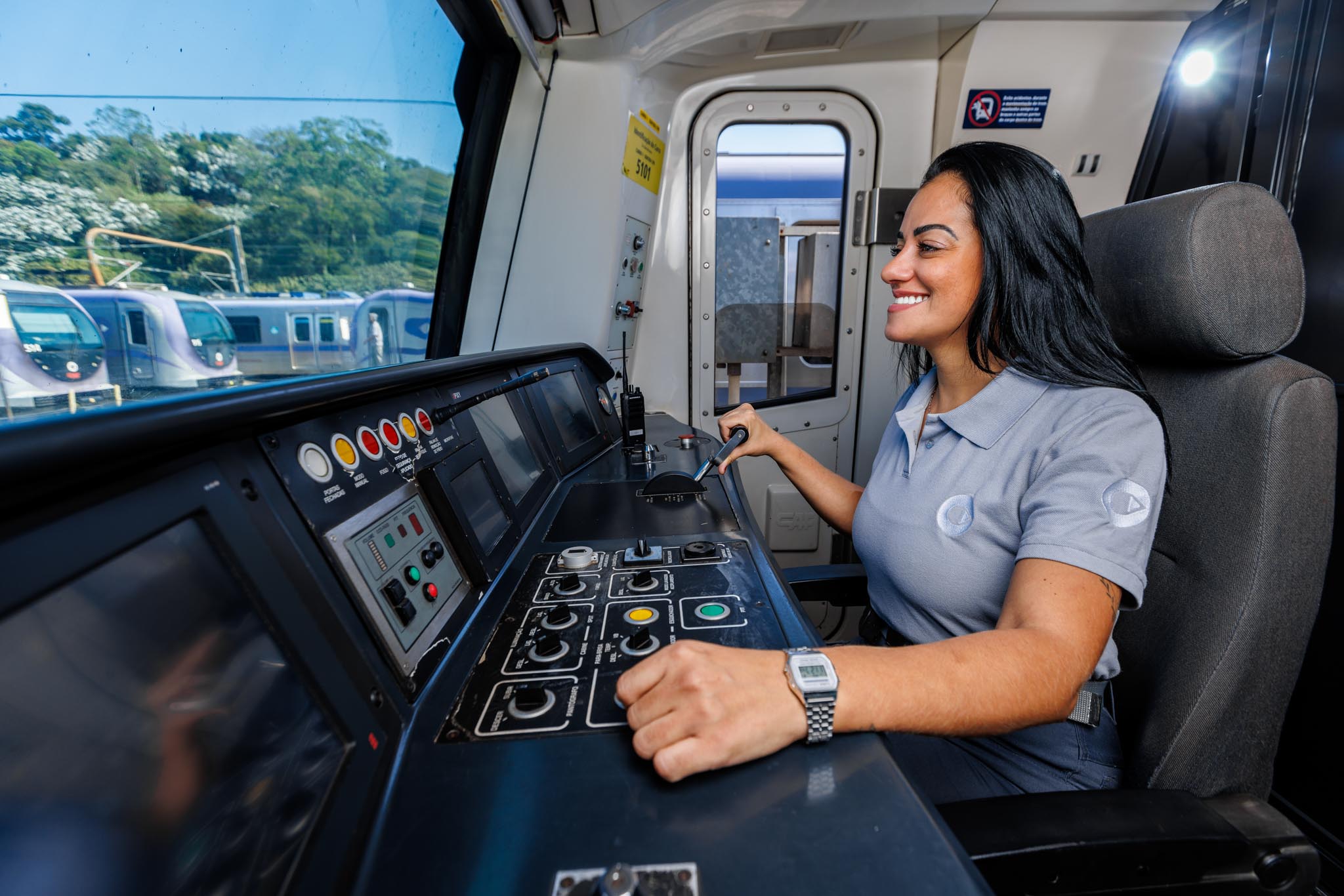 Operadora de trem na cabine de comando, acionando os controles com trens estacionados visíveis pela janela.