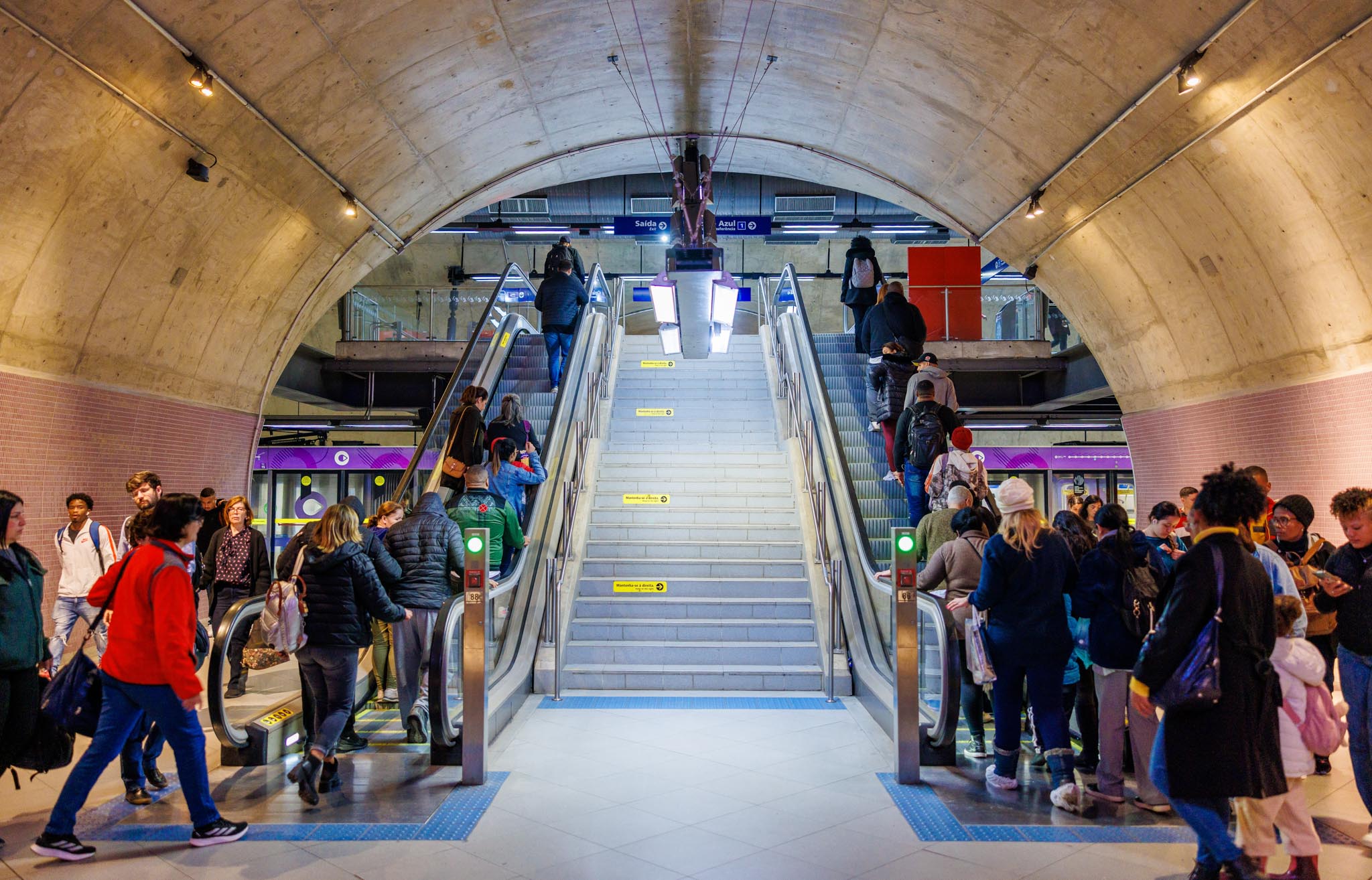 Interior de estação de metrô com escadas rolantes, escada central e muitos passageiros circulando pelo local.